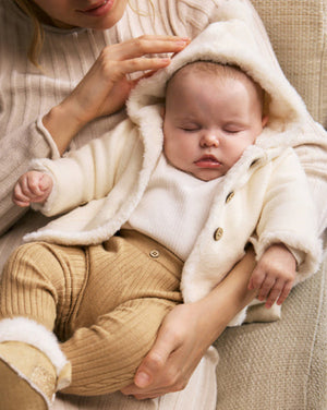 Baby in a white coat being held by a person on a beige couch