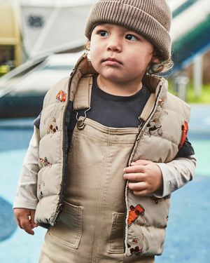 Child wearing a beige puffer vest and overalls with a knit hat, standing outdoors.