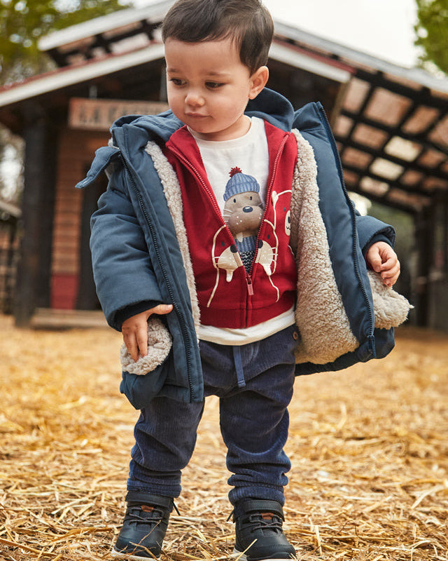 Child wearing a blue coat with a red shirt featuring a bear design, standing on straw.