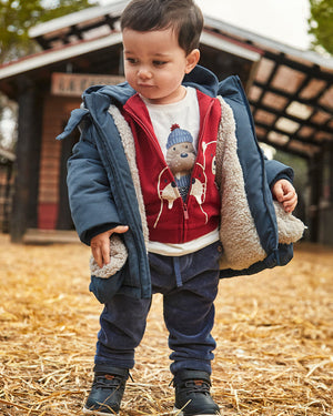 Child wearing a blue coat with a red shirt featuring a bear design, standing on straw.