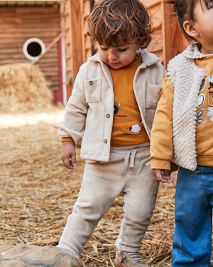 Two children in a barn setting, one wearing a beige jacket and the other in a yellow jacket with a patterned design.