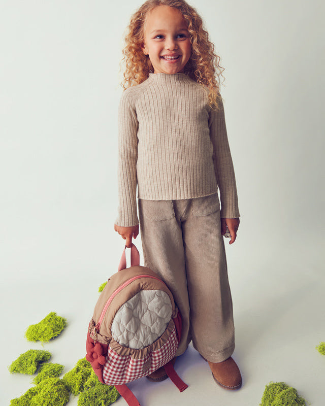 Child wearing a beige sweater and pants with a pink backpack on a light gray background