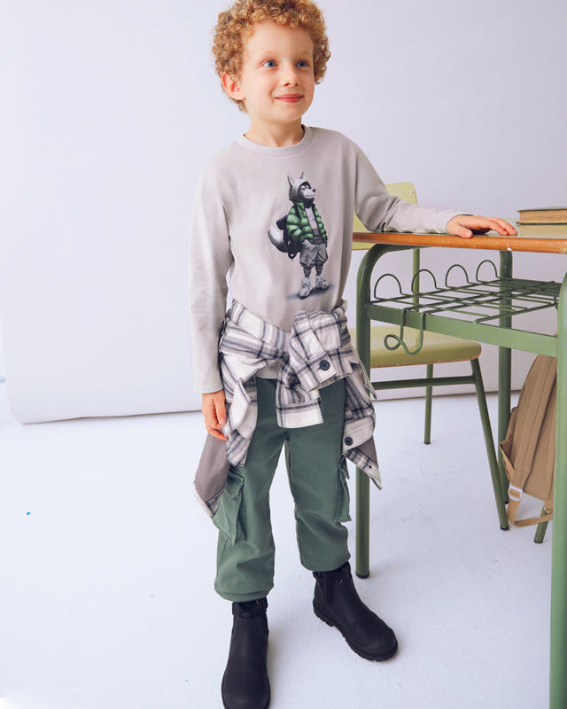 Child wearing a gray sweater with a graphic design, green pants, and black boots in a room with a white wall and wooden table.