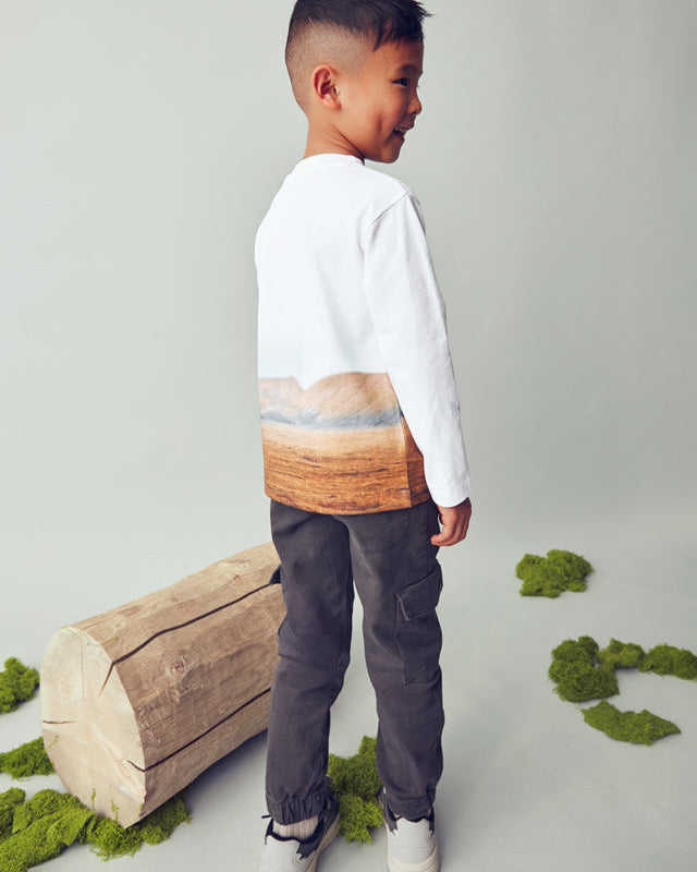 Child wearing a white shirt with a colorful gradient design, standing next to a wooden log and greenery on a light gray background.