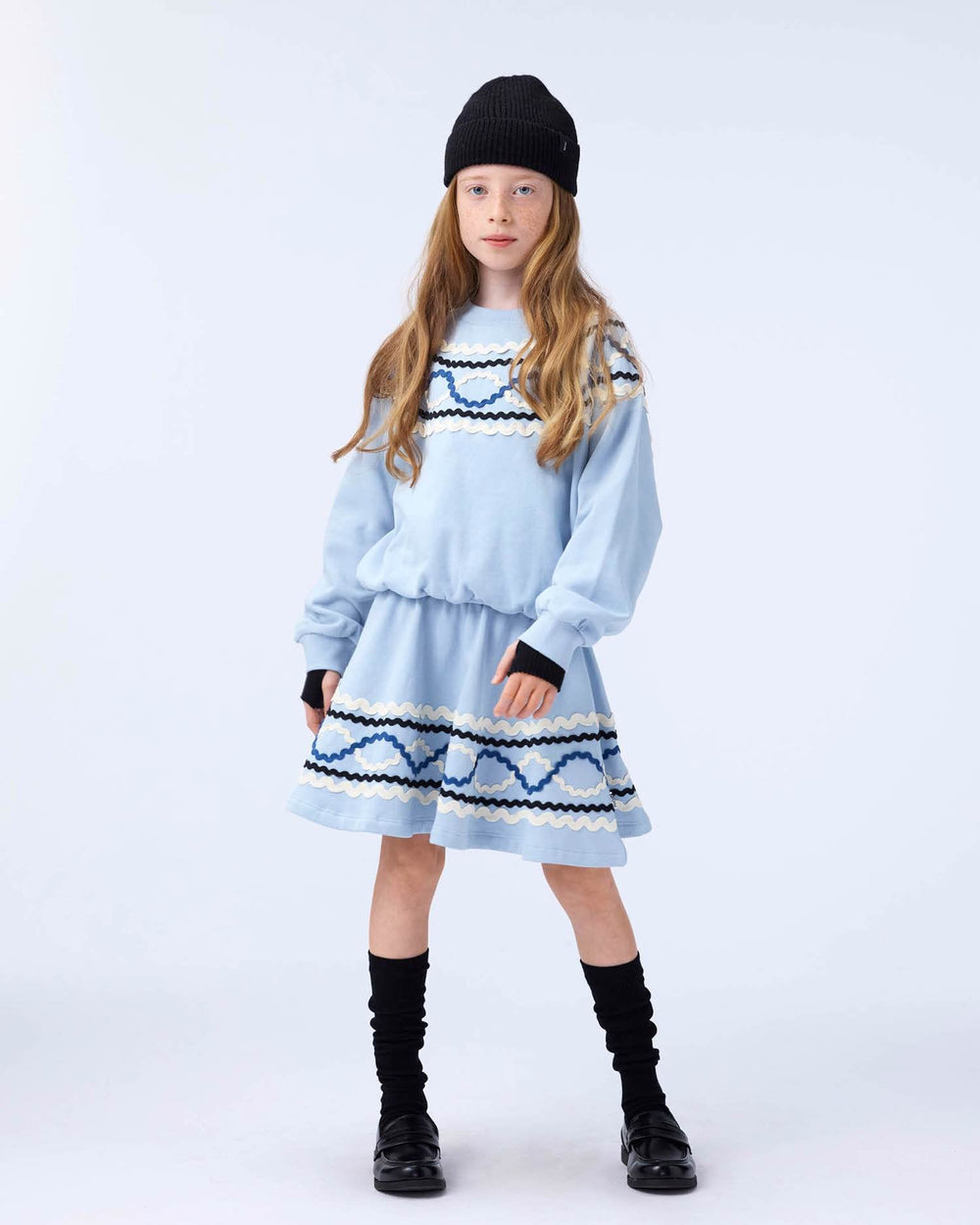 Young girl wearing a light blue dress with black patterns and black boots on a white background