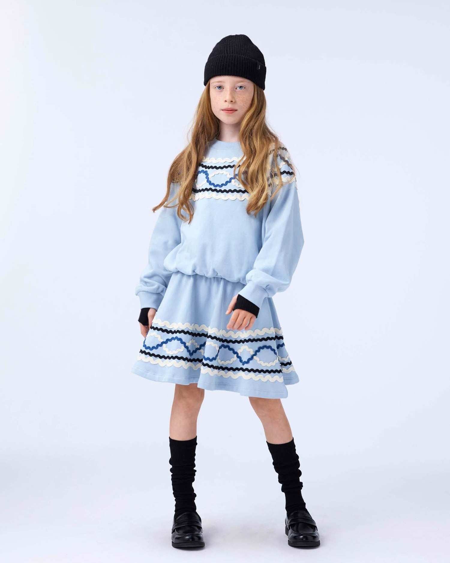 Young girl wearing a light blue dress with black patterns and black boots on a white background