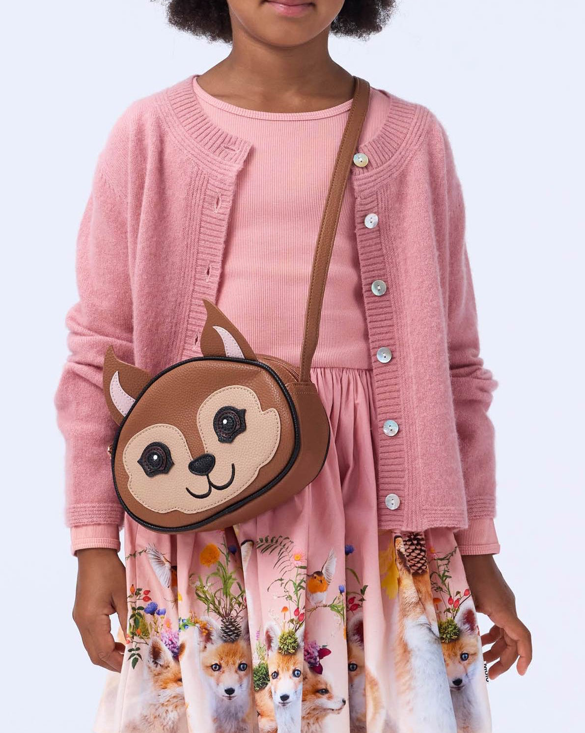Child wearing a pink cardigan and dress with a bear-themed bag on a light blue background