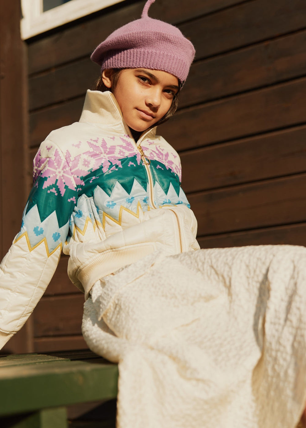 Person wearing a colorful patterned jacket and pink beret sitting against a wooden wall.