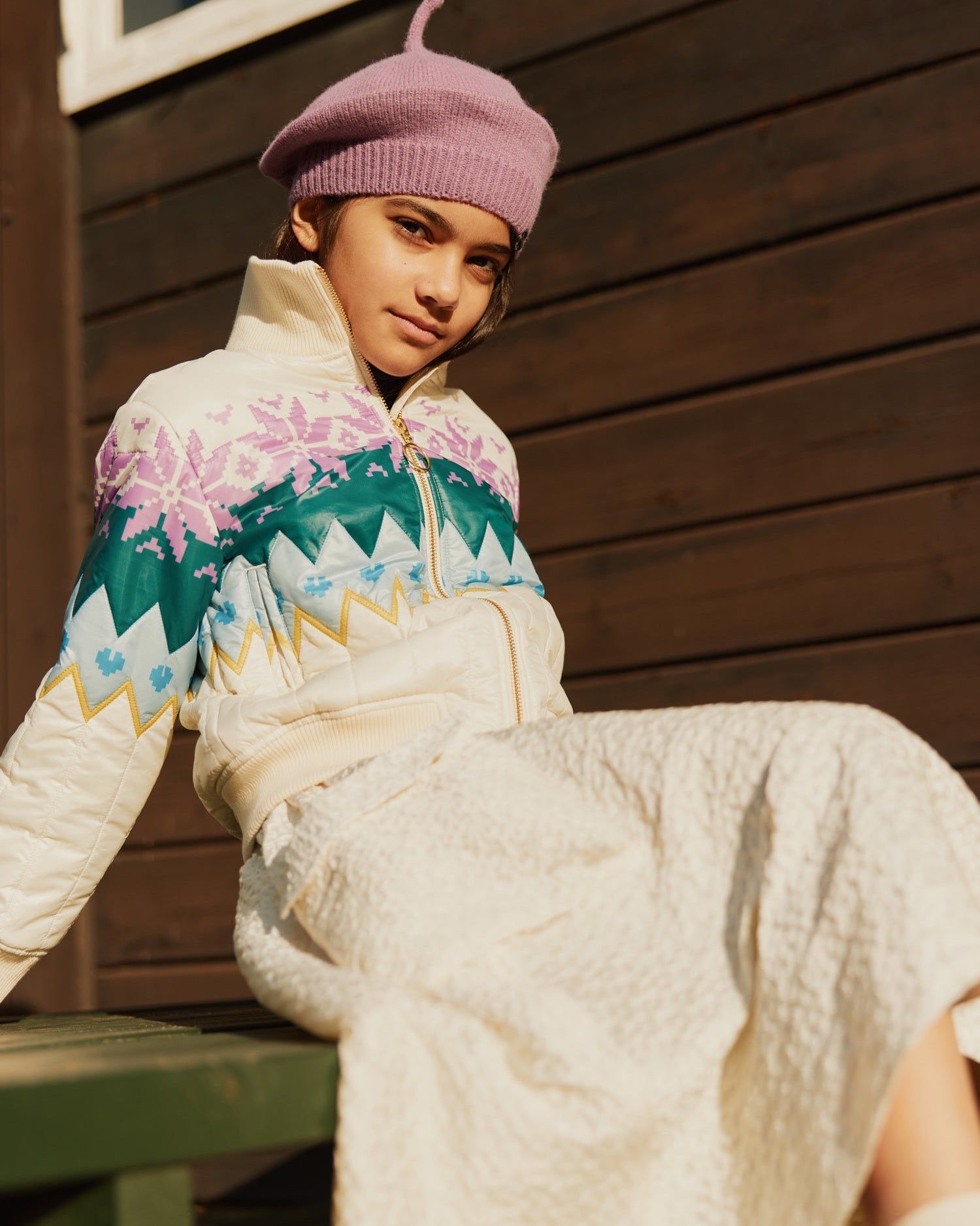 Person wearing a colorful patterned jacket and pink beret sitting against a wooden wall.