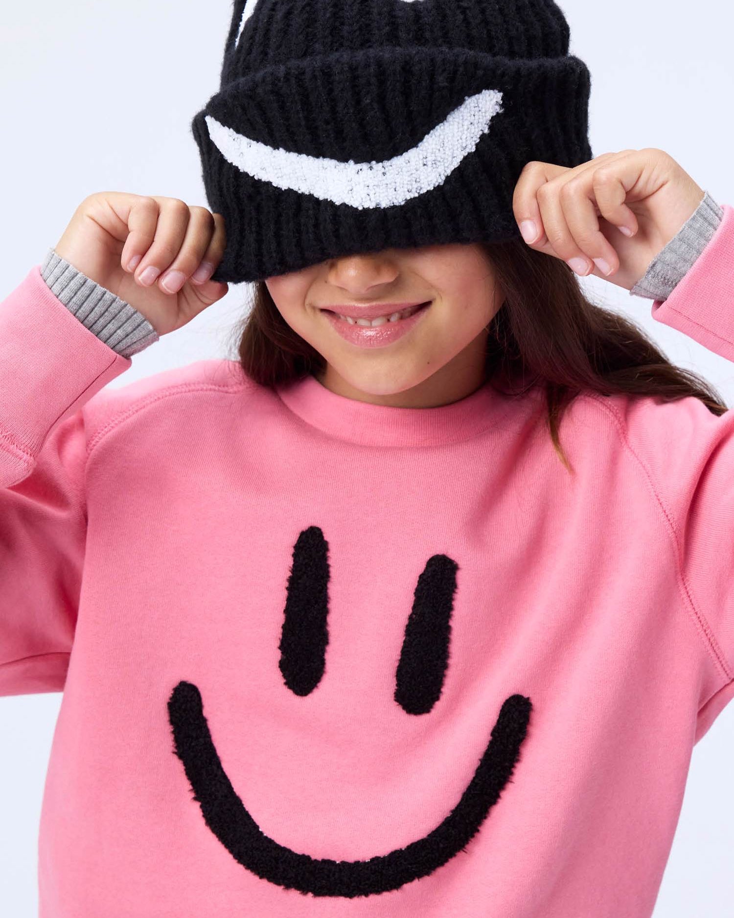 Person wearing a black knit hat with a smiley face and a pink sweater with a smiley face on a light gray background