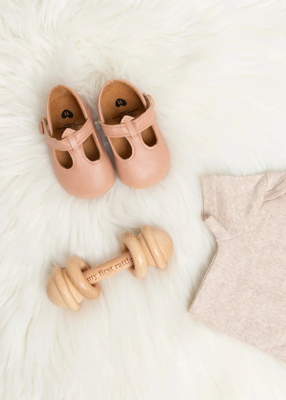 Pink baby shoes, wooden toy, and beige onesie on a white surface