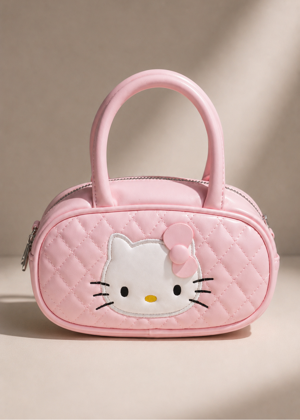 A pink quilted handbag featuring a large Hello Kitty face patch on the front and a matching pink bow, shown on a warm beige background with soft shadows.