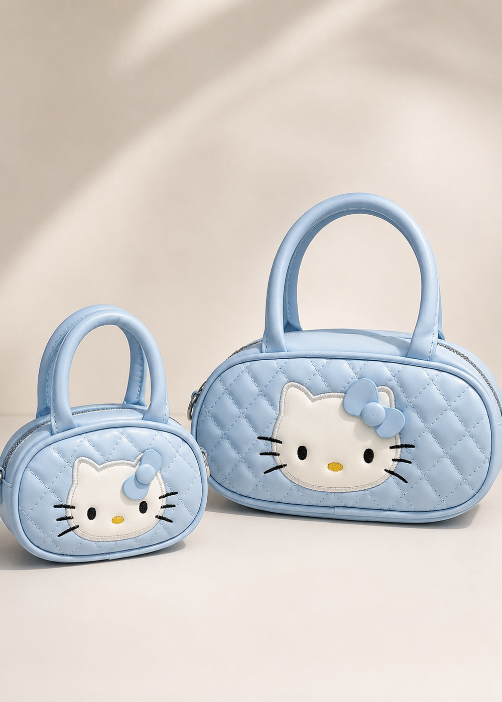 Two light blue quilted handbags with Hello Kitty faces, one small and one large, displayed on a warm beige background with soft shadows.