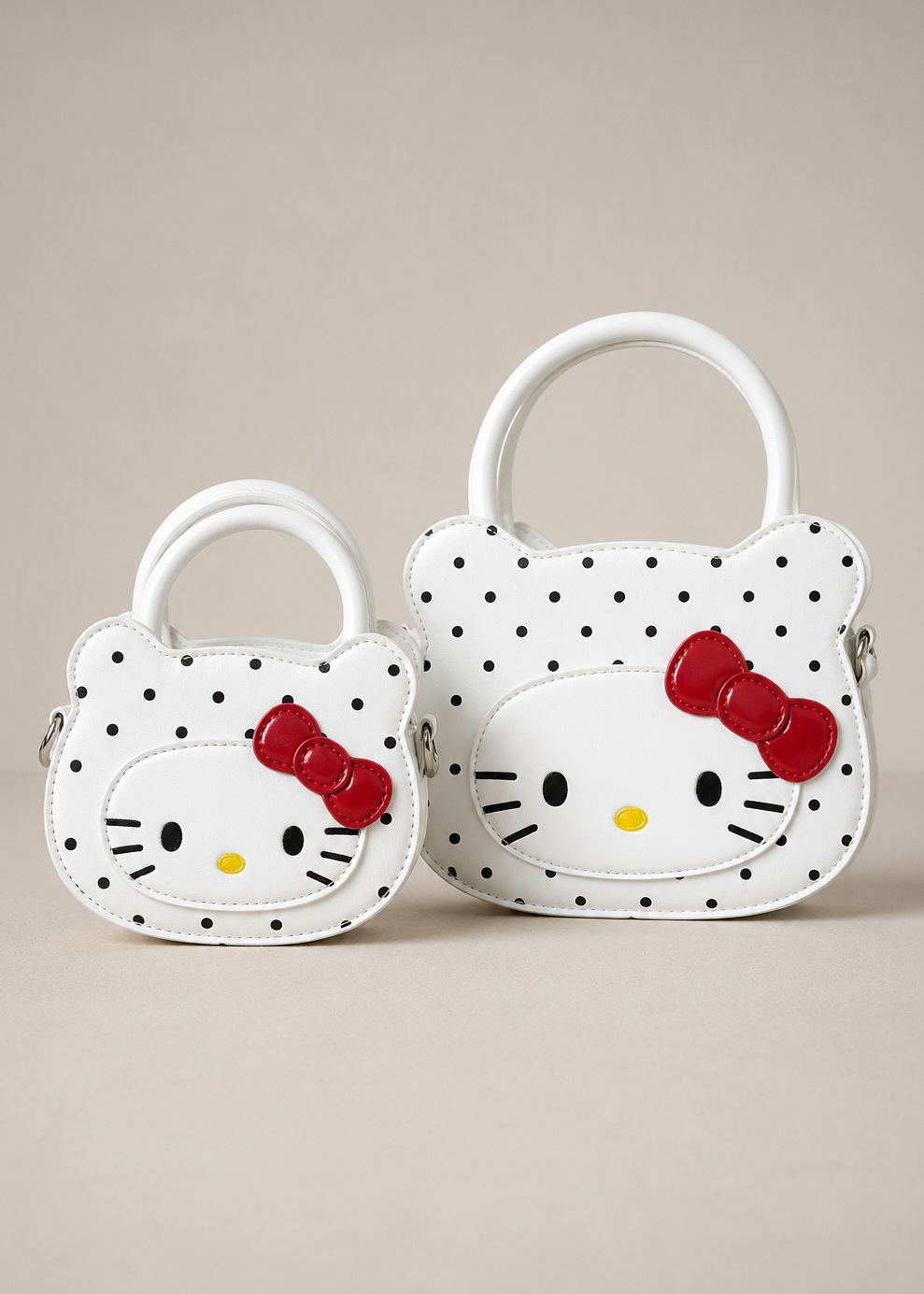 Two small handbags shaped like Hello Kitty's face, one large and one small, in white faux leather with black polka dots, featuring a red bow and Hello Kitty face design, set against a solid beige background.
