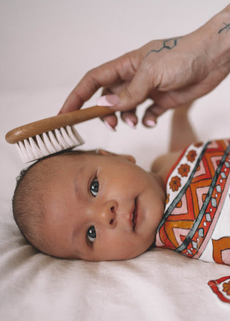 Baby wrapped in a patterned blanket with a hand holding a brush near its head.