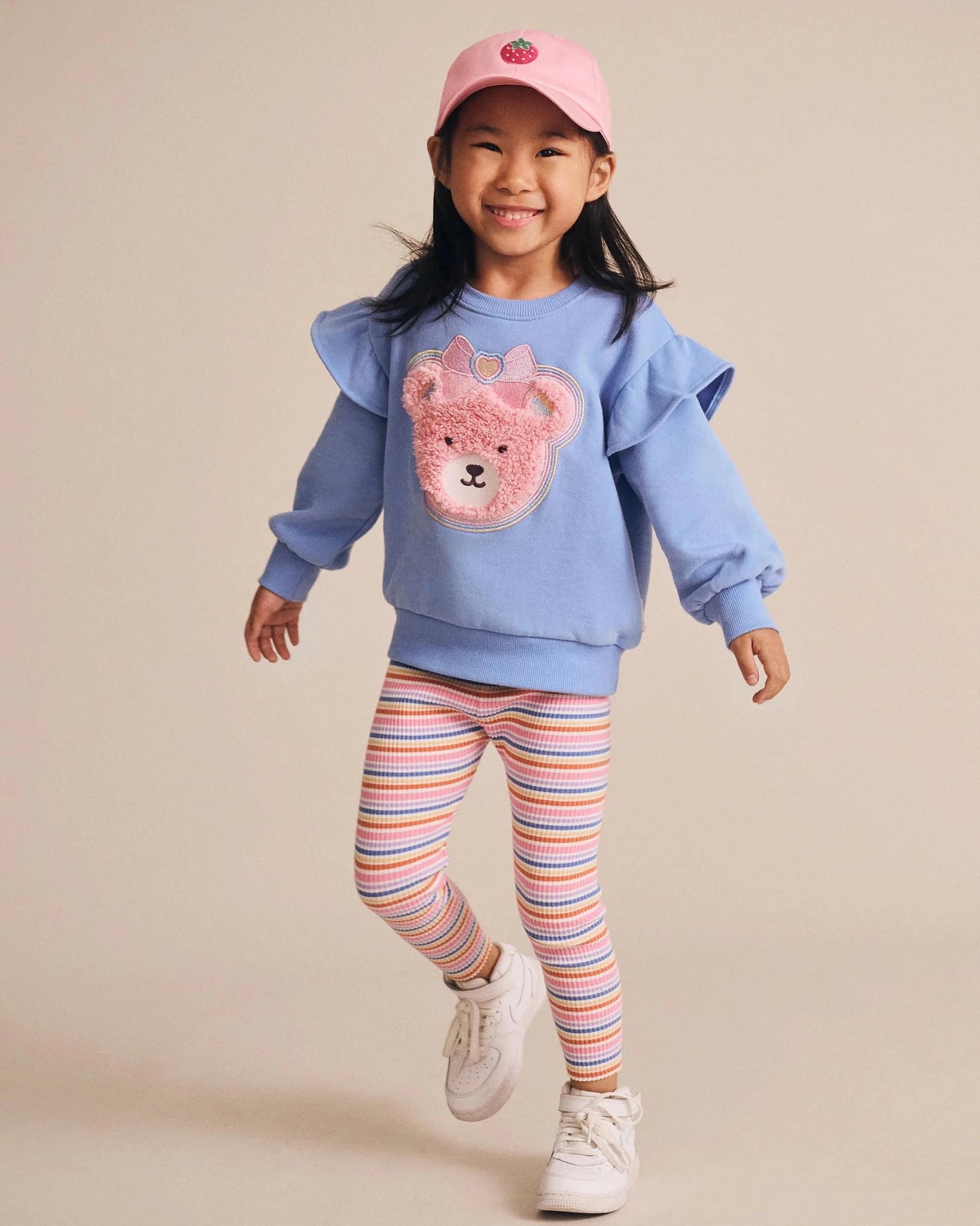 Child wearing a blue sweater with a bear design, striped leggings, and a pink cap on a beige background