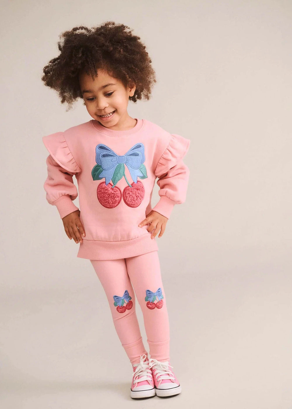 A young girl with curly brown hair wearing a pink ruffle-sleeve sweatshirt and matching leggings with cherry and bow appliques, paired with pink sneakers, against a plain light beige studio background.