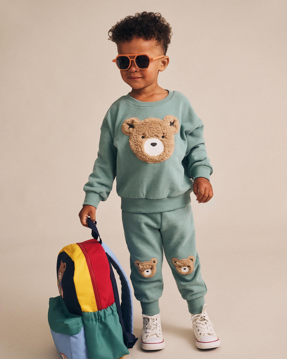 Child wearing a green sweater and pants with bear designs, holding colorful bags.