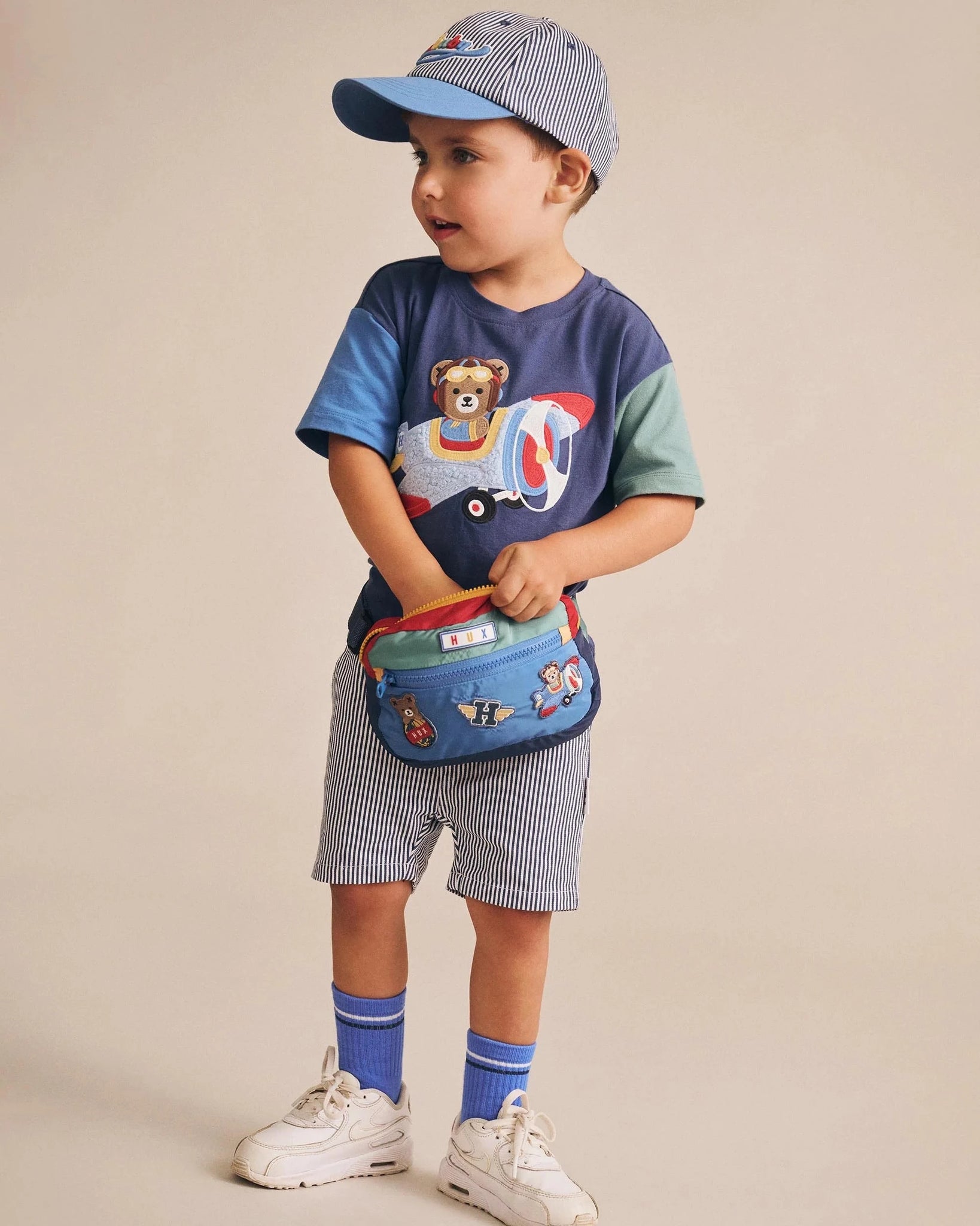 Child wearing a blue shirt with a bear design, shorts, and a cap, holding a colorful bag on a beige background.