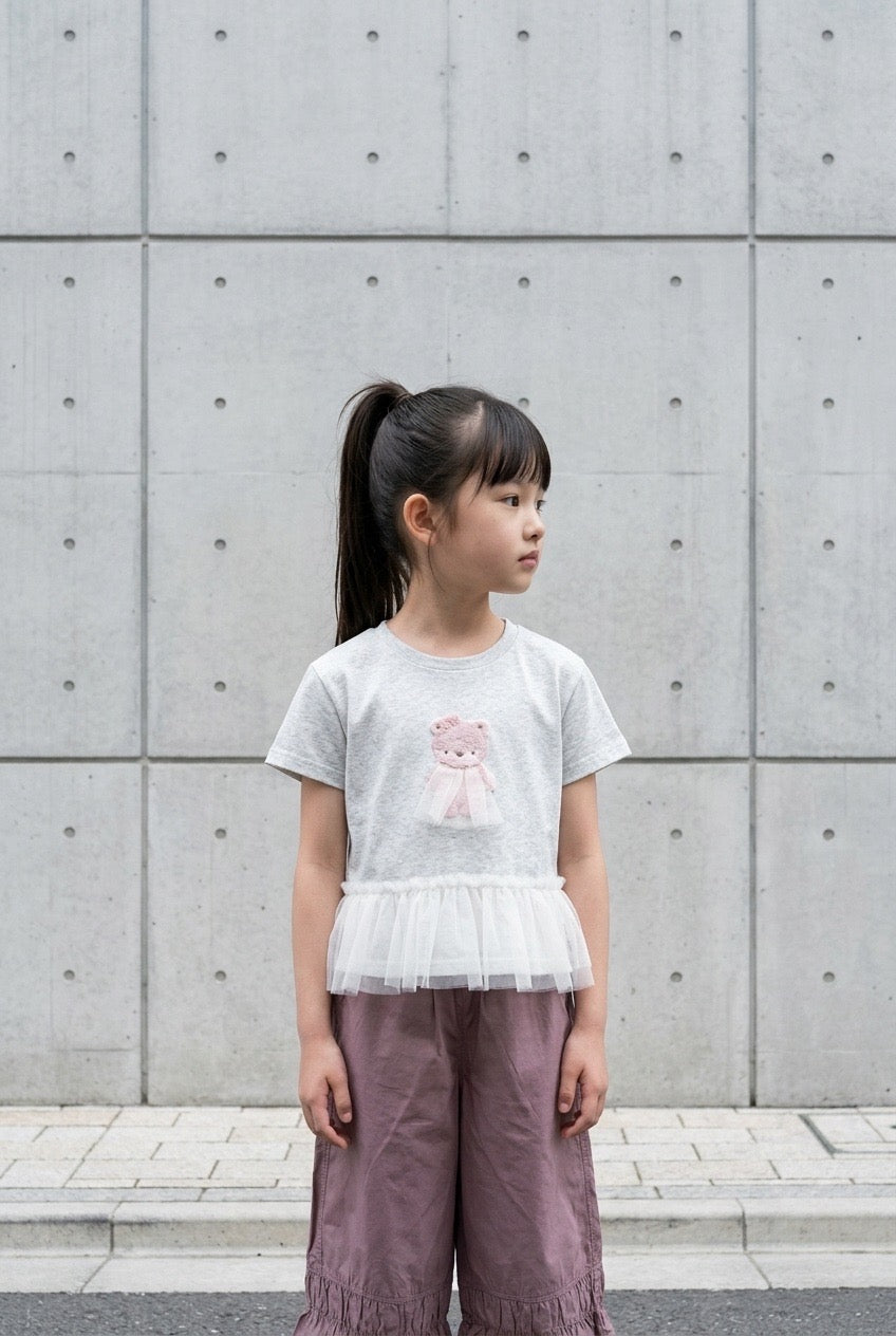 Young girl wearing a light gray t-shirt with a pink design and purple pants standing against a gray concrete wall.