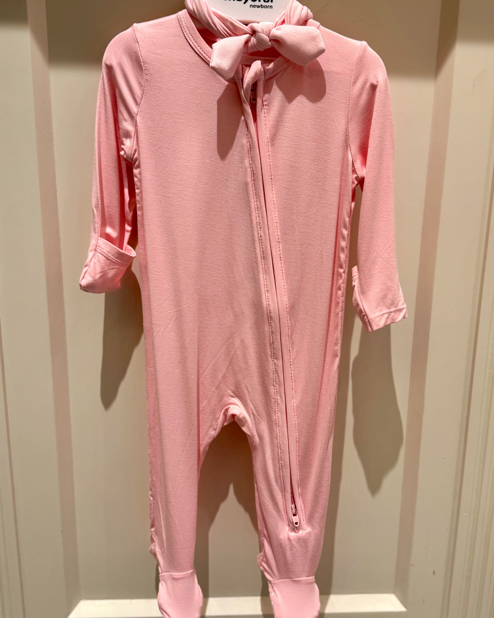 Pink baby onesie with a bow on a hanger against a beige wall.