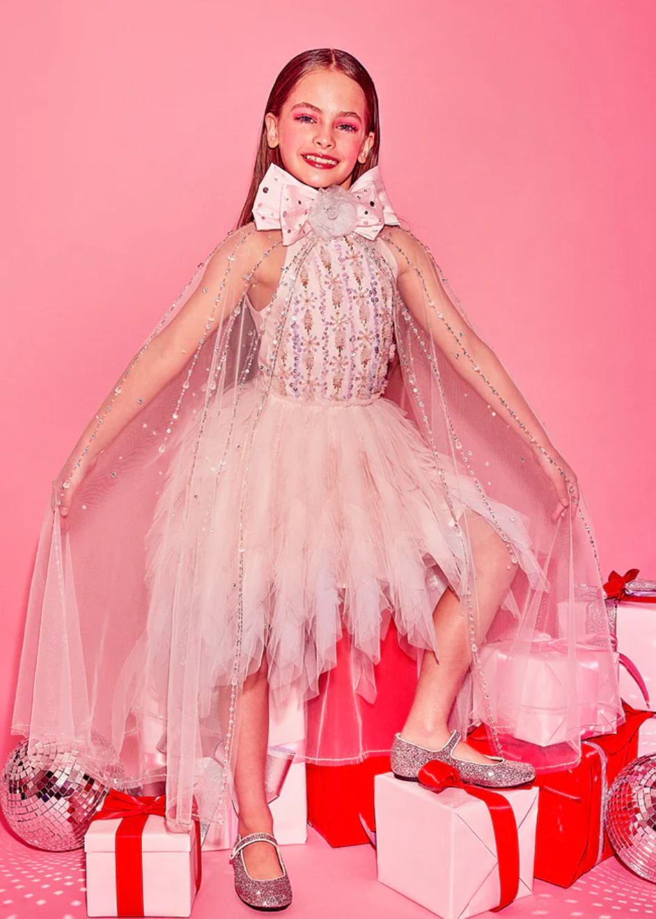 A girl wearing a pink tutu dress with sequins and floral embellishments, posing on a set with Christmas-themed decorations including presents and a mirrored ball.