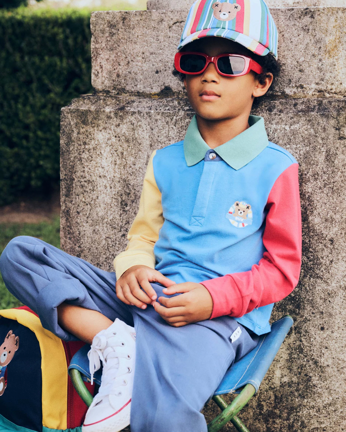Child wearing a colorful outfit with a vest and sunglasses, sitting on a stone bench outdoors.