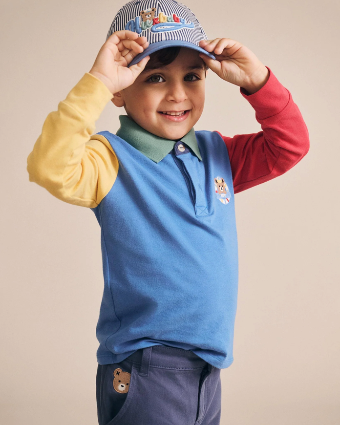 Child wearing a colorful outfit with a blue vest and cap, standing against a beige background