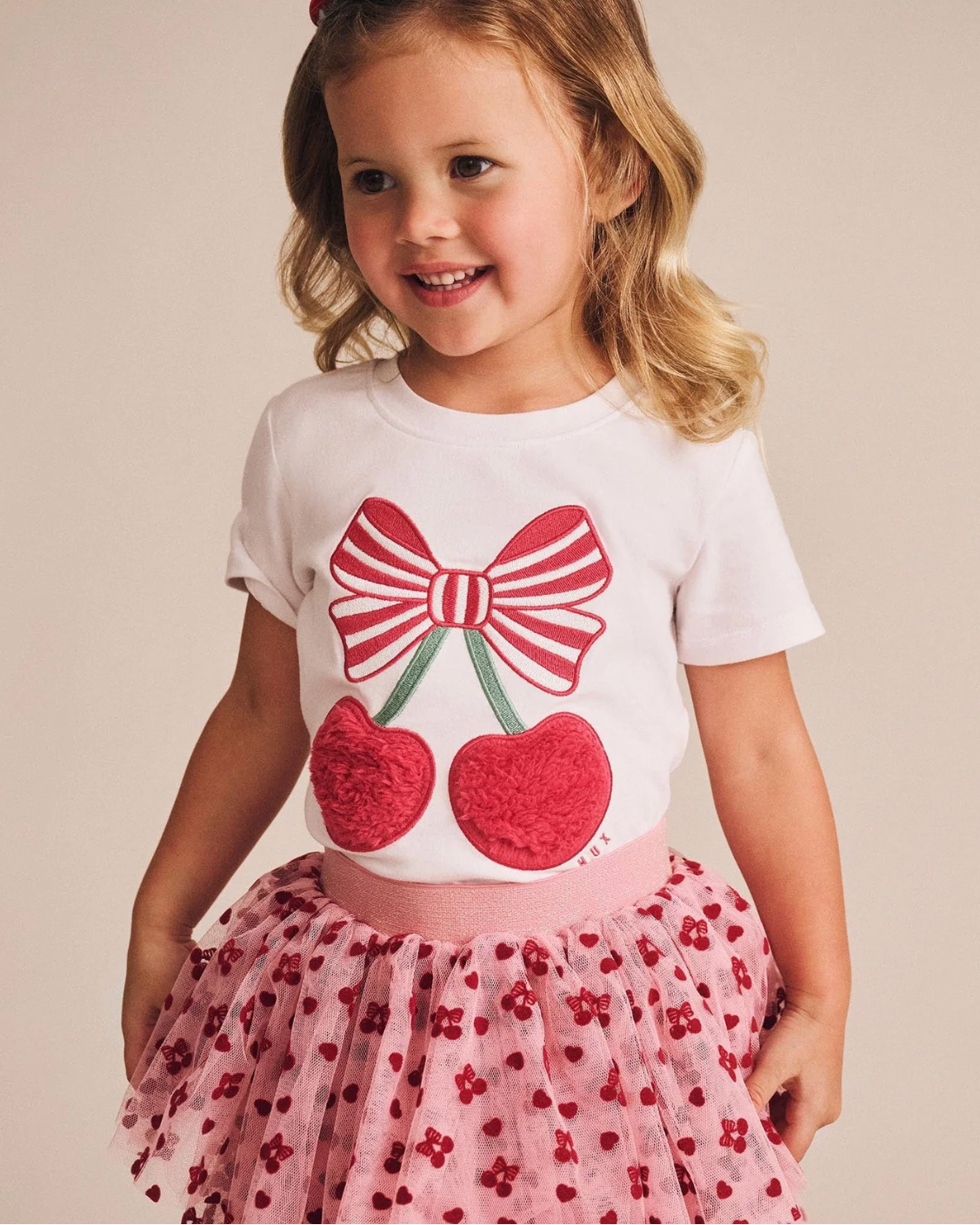 Young girl wearing a white t-shirt with a red bow and cherry design, paired with a pink skirt.