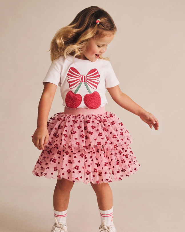 Young girl wearing a white t-shirt with a red bow and cherries design, paired with a pink floral skirt on a beige background.