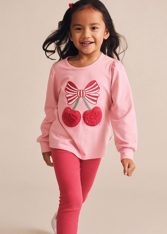Young girl wearing a pink sweater with a bow and cherry design and pink leggings on a beige background.