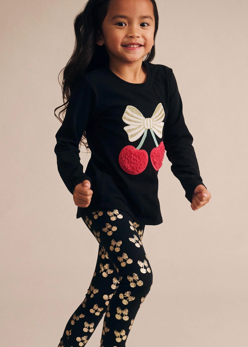 Child wearing a black sweater with a bow and cherry design, paired with black pants with gold patterns on a beige background.