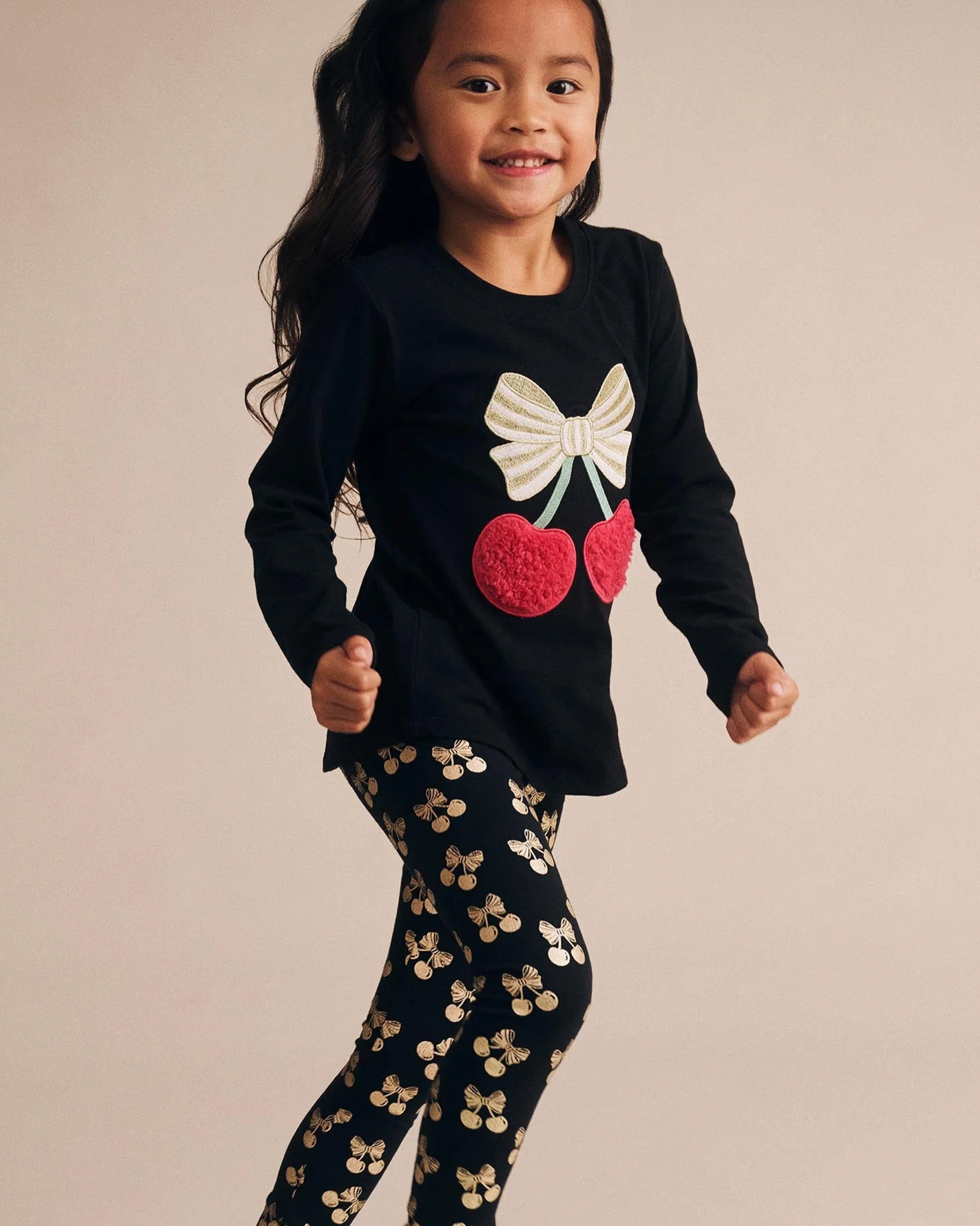 Child wearing a black sweater with a bow and cherry design, paired with black pants with gold patterns on a beige background.