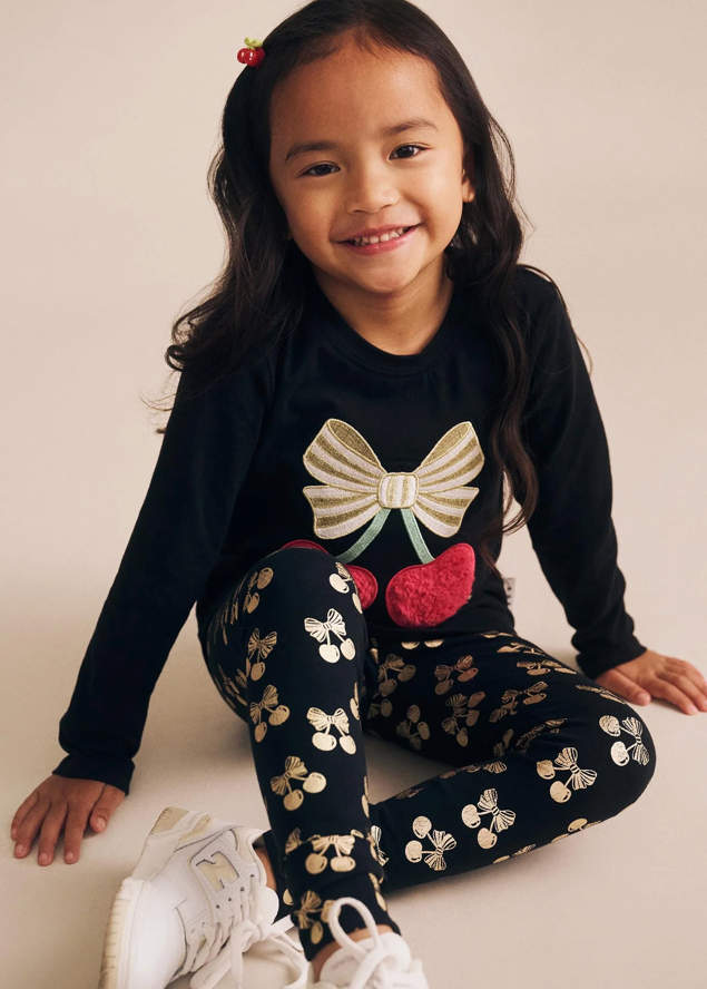 Young girl wearing a black long-sleeve top with a bow design and patterned leggings, sitting on a light-colored floor.