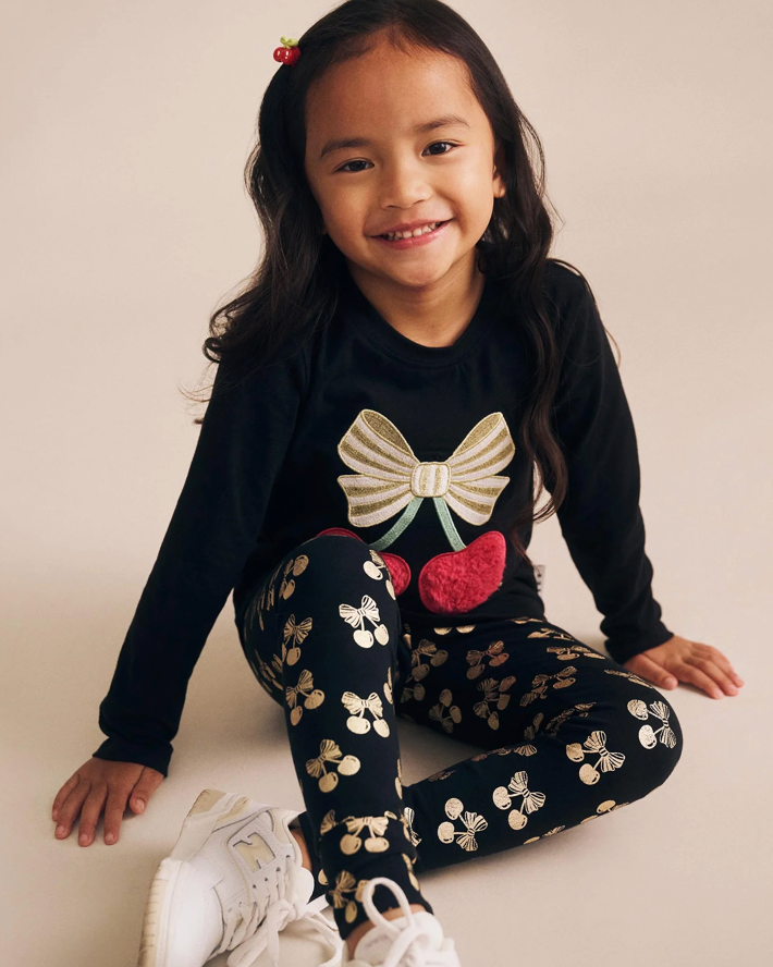Young girl wearing a black long-sleeve top with a bow design and patterned leggings, sitting on a light-colored floor.
