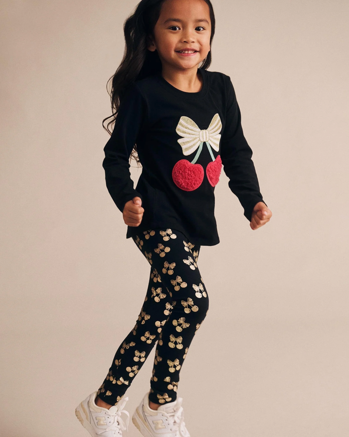 Child wearing a black long-sleeve shirt with a bow and cherry design and black leggings with white patterns on a beige background.