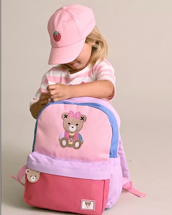 Child wearing a pink backpack and cap with a bear design on a beige background