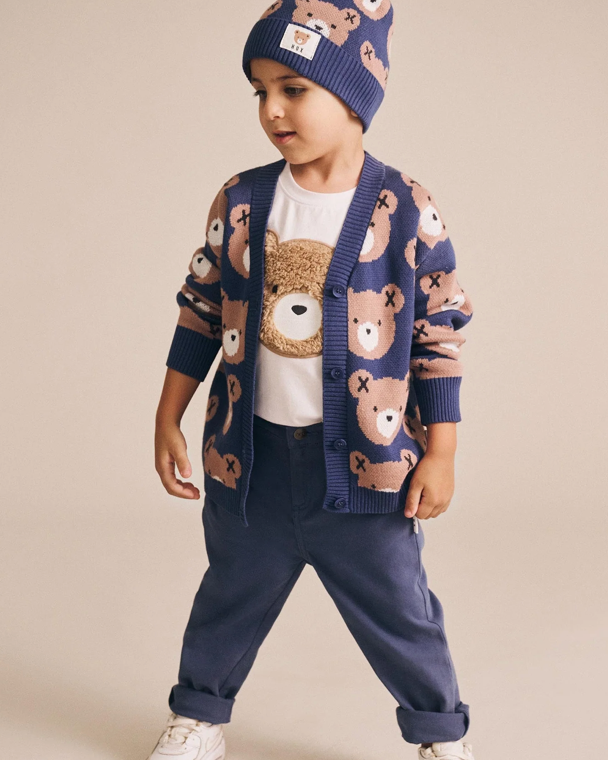 Child wearing a navy cardigan with teddy bear pattern, white shirt, blue pants, and matching hat on a beige background.