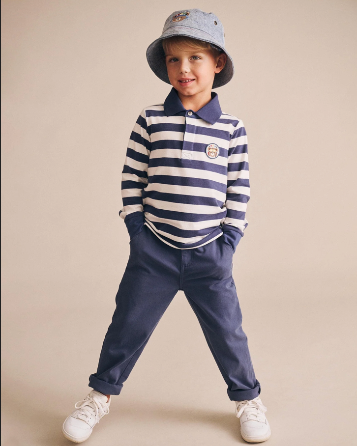 Child wearing a striped polo shirt, navy pants, and a bucket hat on a beige background