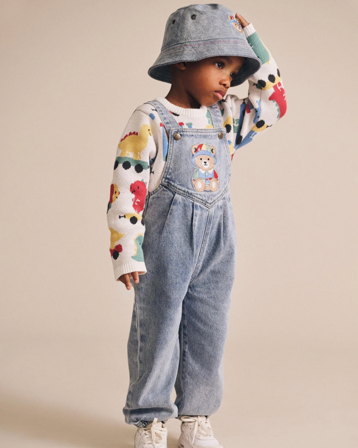 Child wearing a colorful sweater, denim overalls, and a gray bucket hat on a beige background