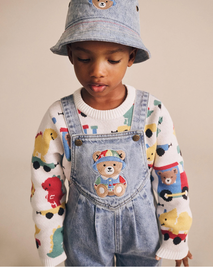 Child wearing a denim bucket hat and overalls with teddy bear designs on a beige background