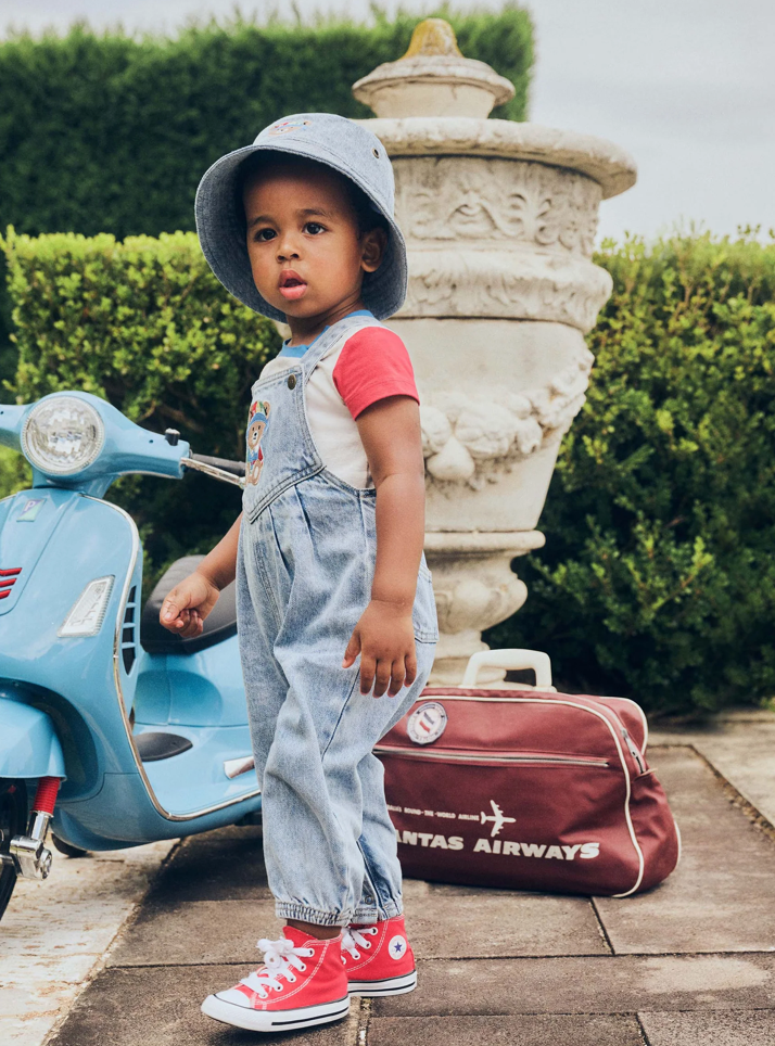 Child in overalls and red shoes standing next to a Vespa scooter and vintage suitcase.