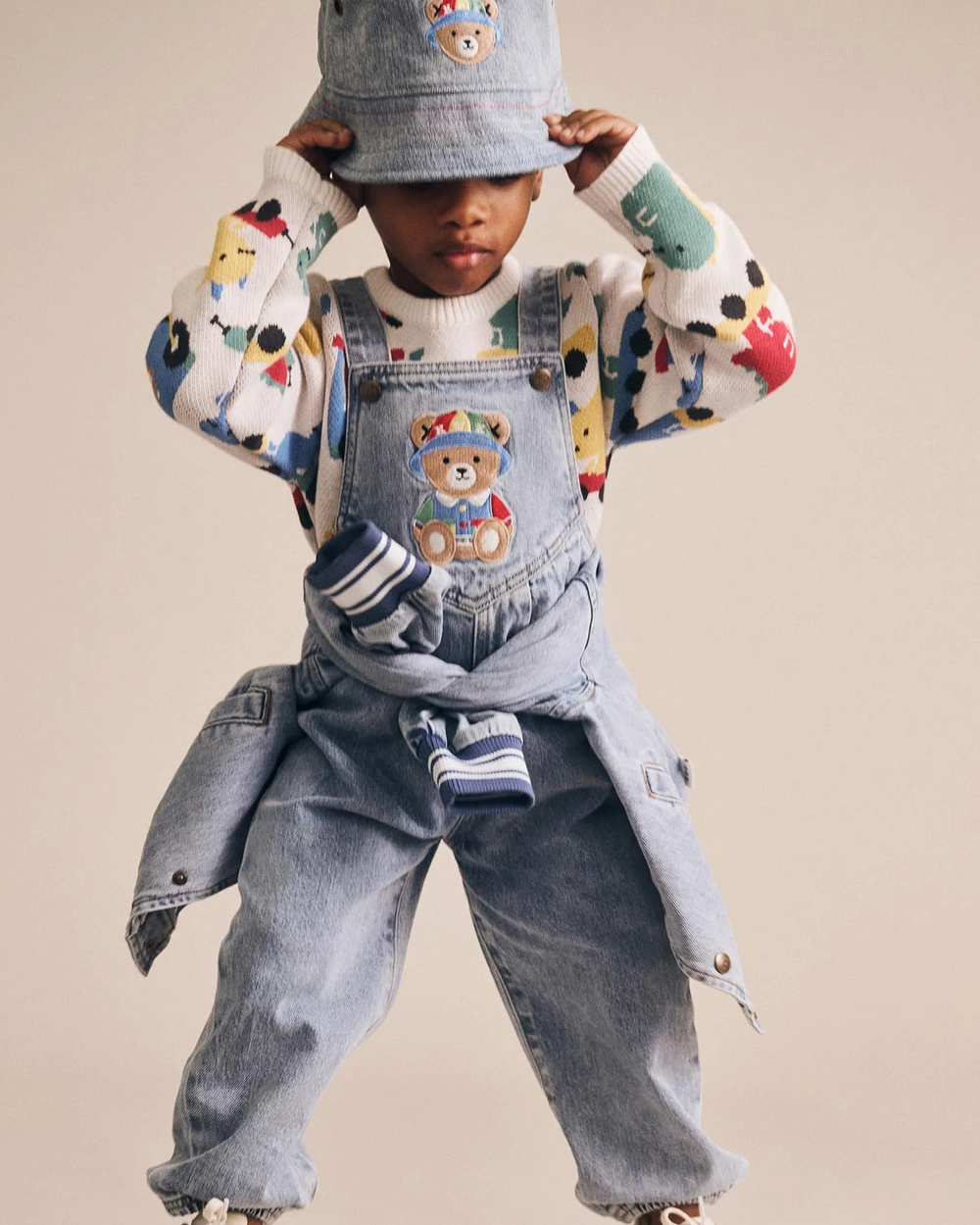 Child wearing a colorful sweater, denim overalls, and a bucket hat with teddy bear designs on a beige background.