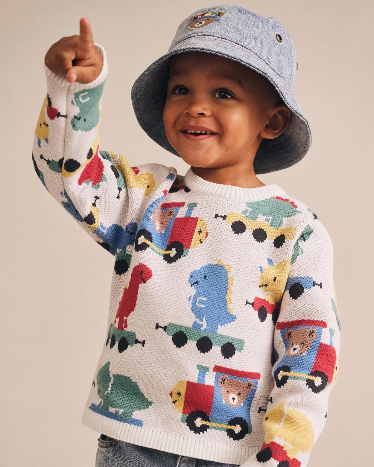 Child wearing a sweater with train designs and a matching hat on a plain background