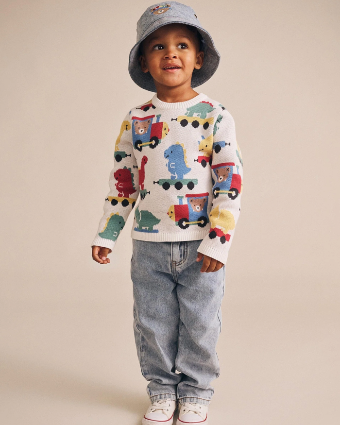 Child wearing a sweater with train designs, jeans, and a bucket hat on a beige background