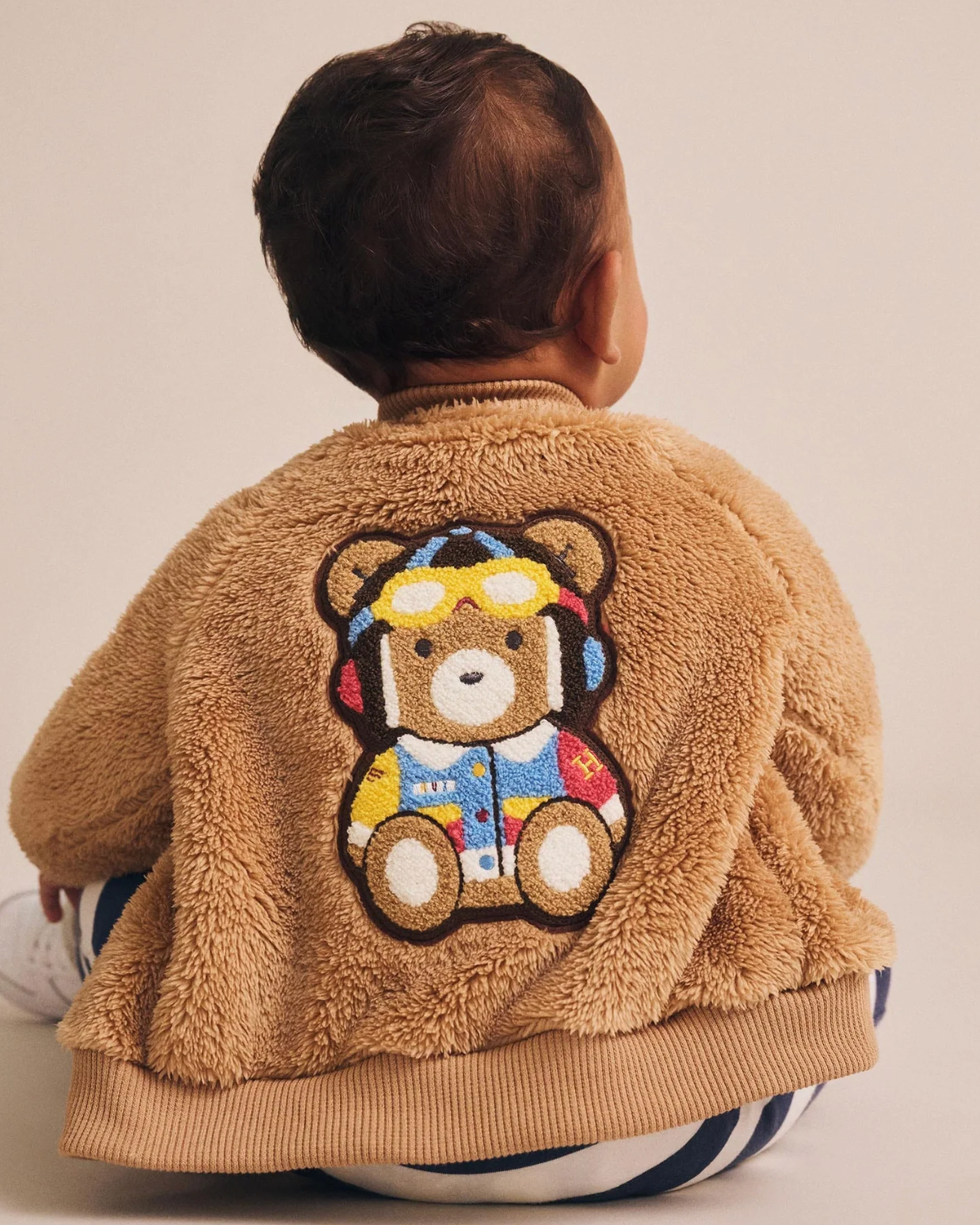 Child wearing a brown teddy bear jacket with a colorful teddy bear design on the back.