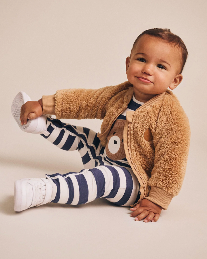 Baby wearing a brown fleece jacket with a teddy bear design, blue and white striped pants, and white shoes on a beige background.