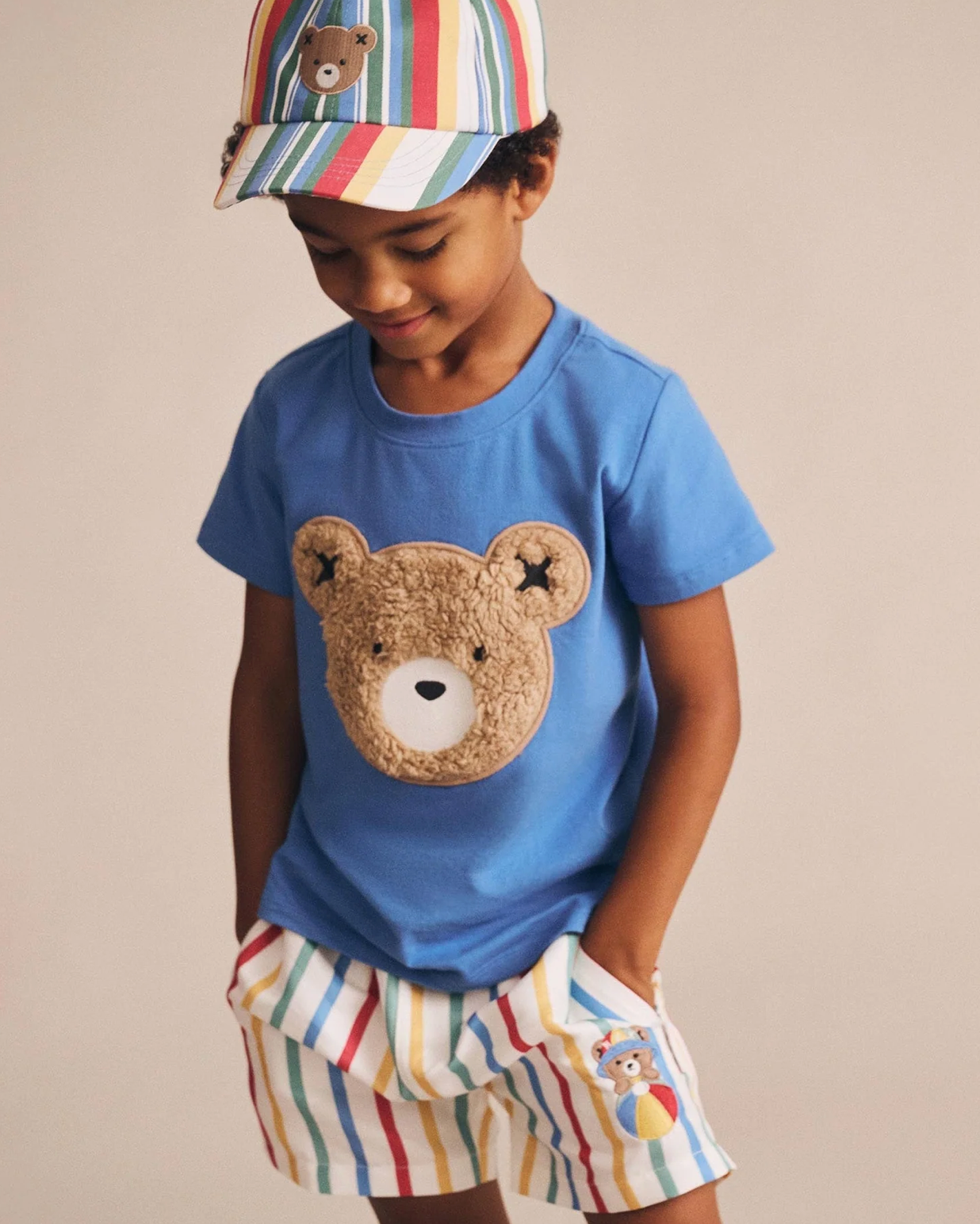 Child wearing a blue t-shirt with a teddy bear design and colorful striped shorts on a beige background