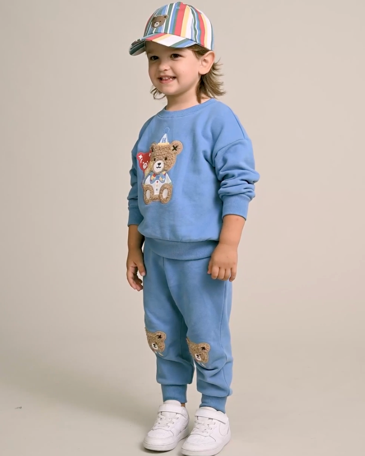 Child wearing a blue outfit with teddy bear designs and a colorful cap on a beige background