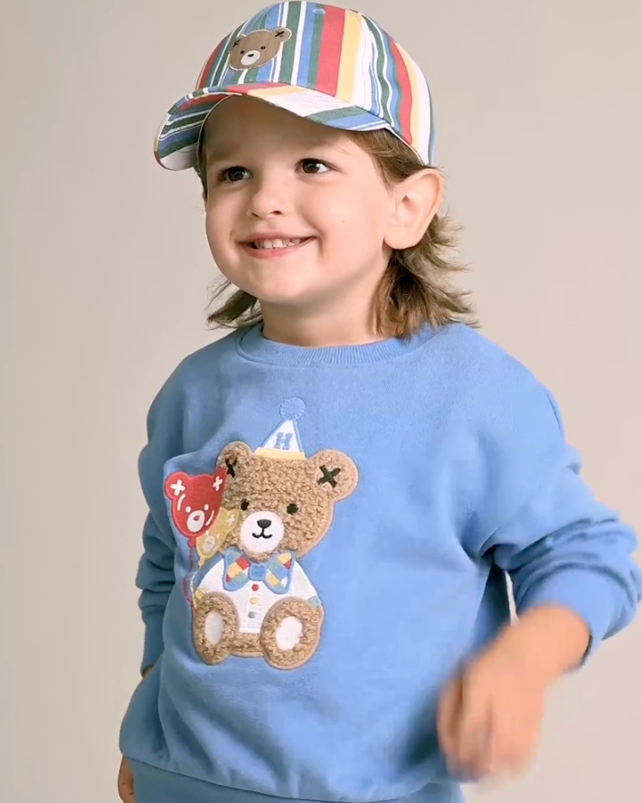 Child wearing a blue sweatshirt with a teddy bear design and a colorful striped cap.
